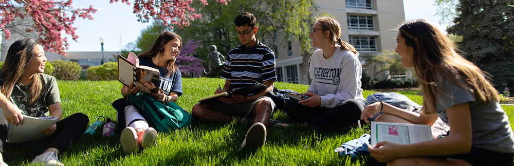 A group of Creighton students sitting outside on the grass while reading books and talking