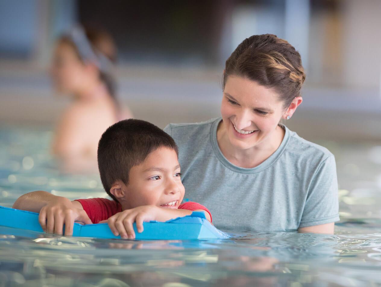 A physical therapist guides a young boy on a flotation device during aquatic exercises