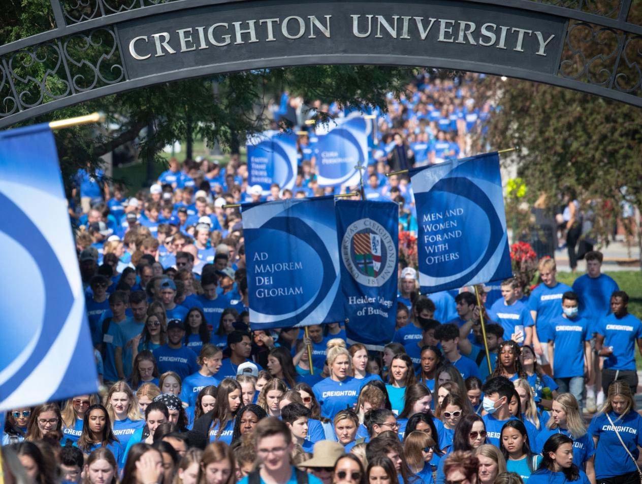 Students walking in mass under Creighton University gate -- all wearing Creighton blue t-shirts.