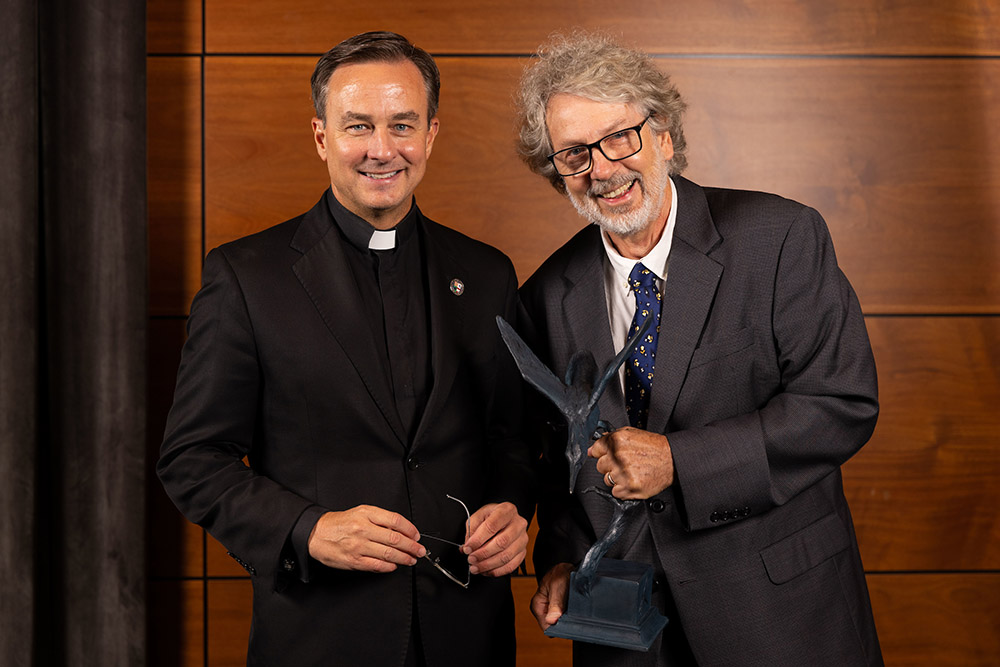 Rev. Daniel S. Hendrickson, SJ, PhD, president of Creighton University, stands at left in black suit with clerical collar; Patrick Murray leans in from right in a gray jacket, holding a life-sized sculpture of a kingfisher (bird) and dragonfly by a branch, representing an image in the poem "As Kingfisher Catch Fire, Dragonflies Draw Flame."