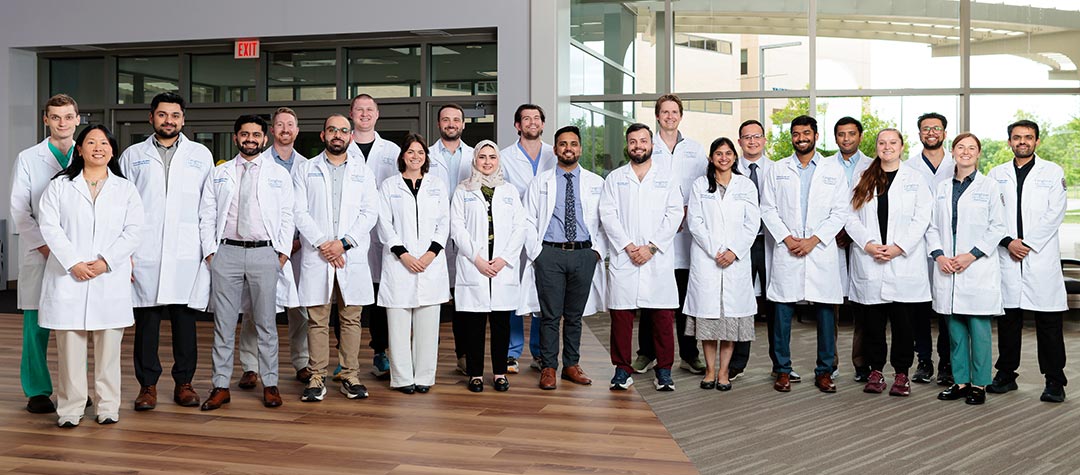 Third-year Internal Medicine residents from the Class of 2026 stand in two rows wearing Creighton University lab coats in a bright lobby with floor-to-ceiling windows and wood flooring.