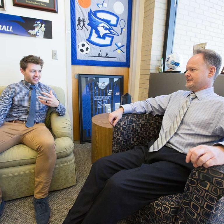 Two male students in business attire sitting in armchairs and conversing