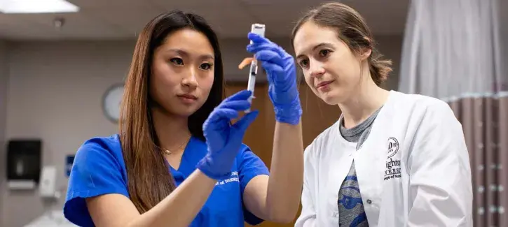 Nurse measuring serum in vial while instructor looks on in white coat.