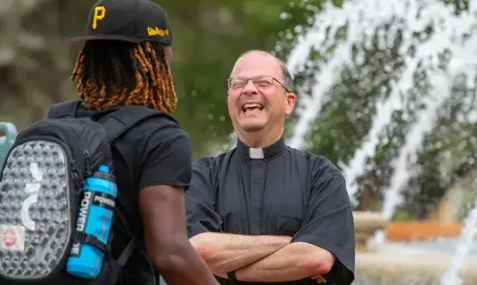 Jesuit interacting with student near fountain.