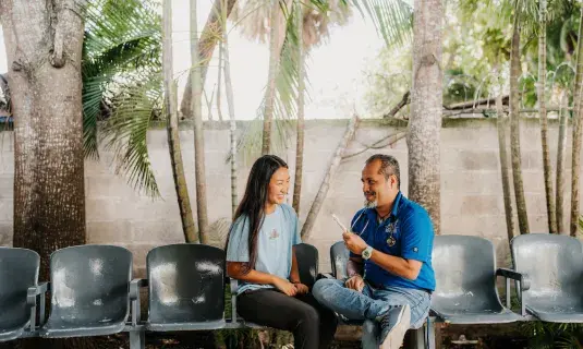 Man an woman sitting on chairs with palm trees in background. 