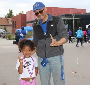 A young girl wearing a medal poses with a peace sign alongside a community health worker at an outdoor event.