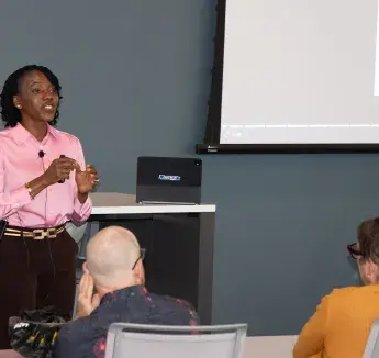 A presenter in a pink shirt leads a health education session in a classroom setting with a projection screen.