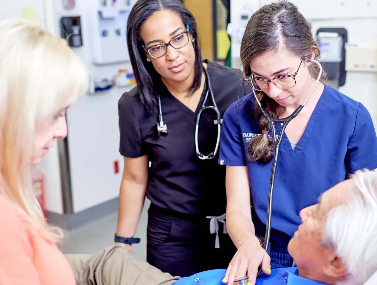 Student practicing using stethoscope on older patient with others standing around.