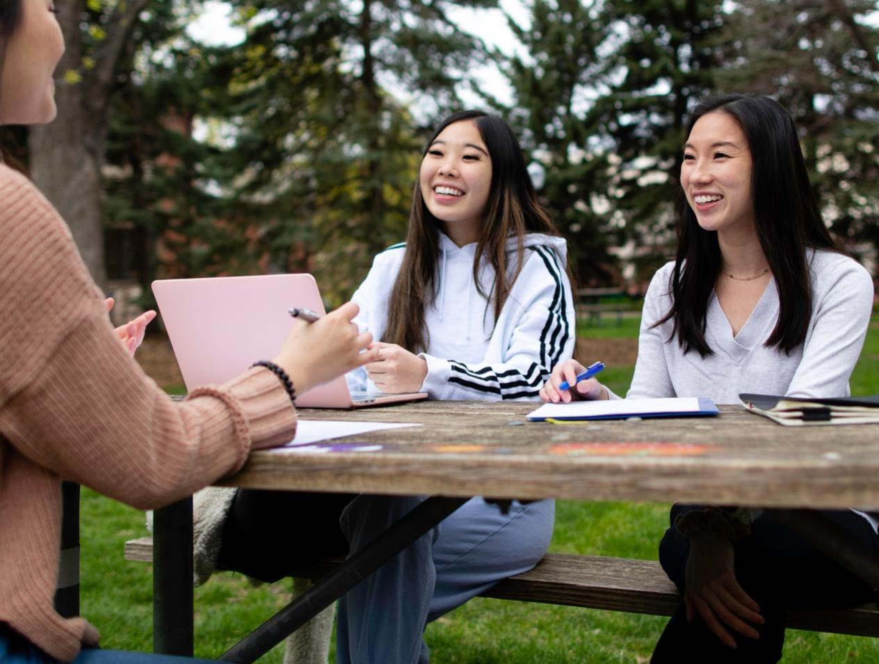 Three women sitting at table outdoors smiling and studying.