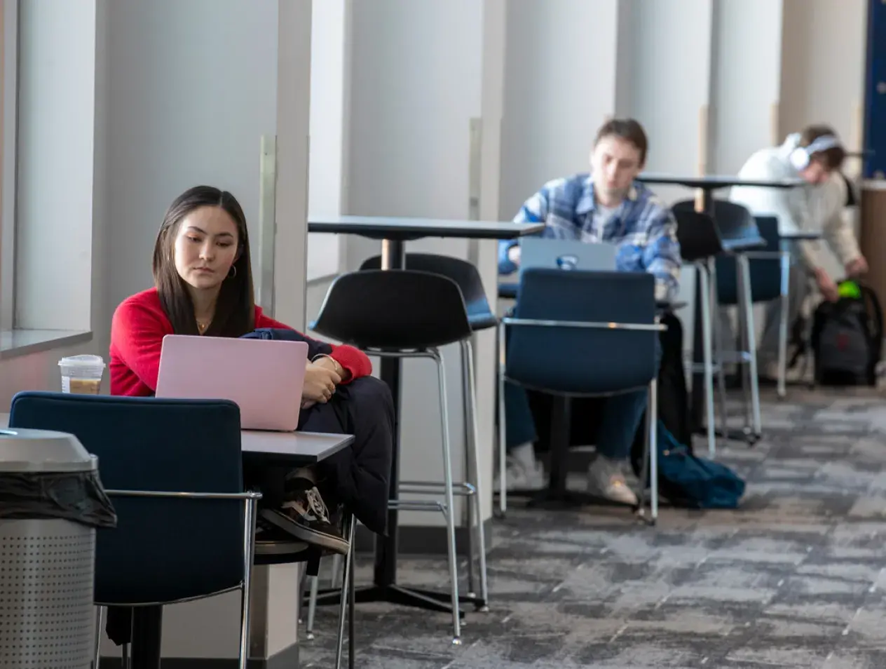 Three students studying individually at tables along a windowed wall