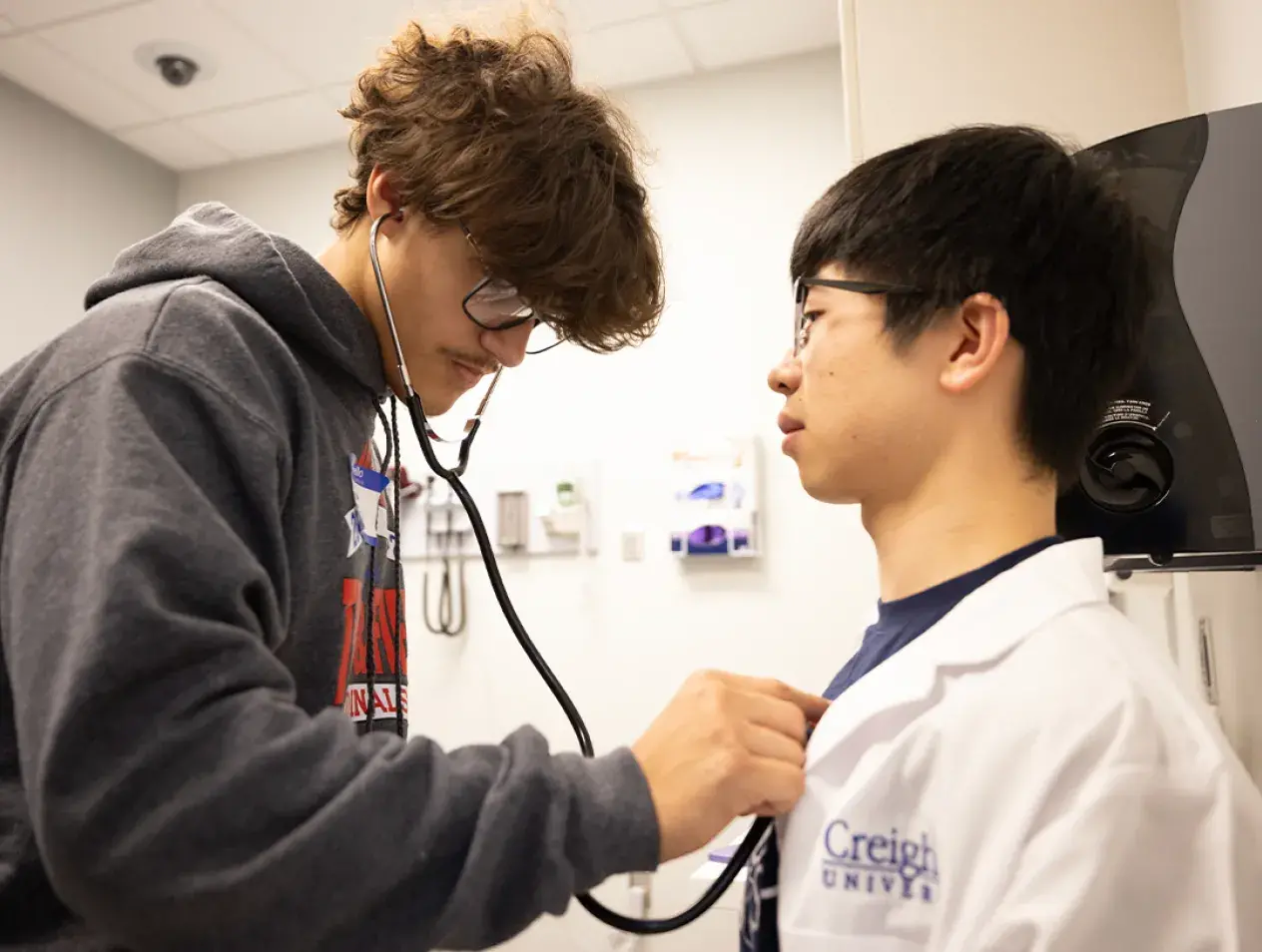The image shows two young men in a clinical environment. One, dressed in a dark hoodie, is using a stethoscope to listen to the other’s chest. The individual being examined wears a white coat with the "Creighton University" logo, indicating a medical education setting. This appears to be part of an educational event, likely a mini medical school.