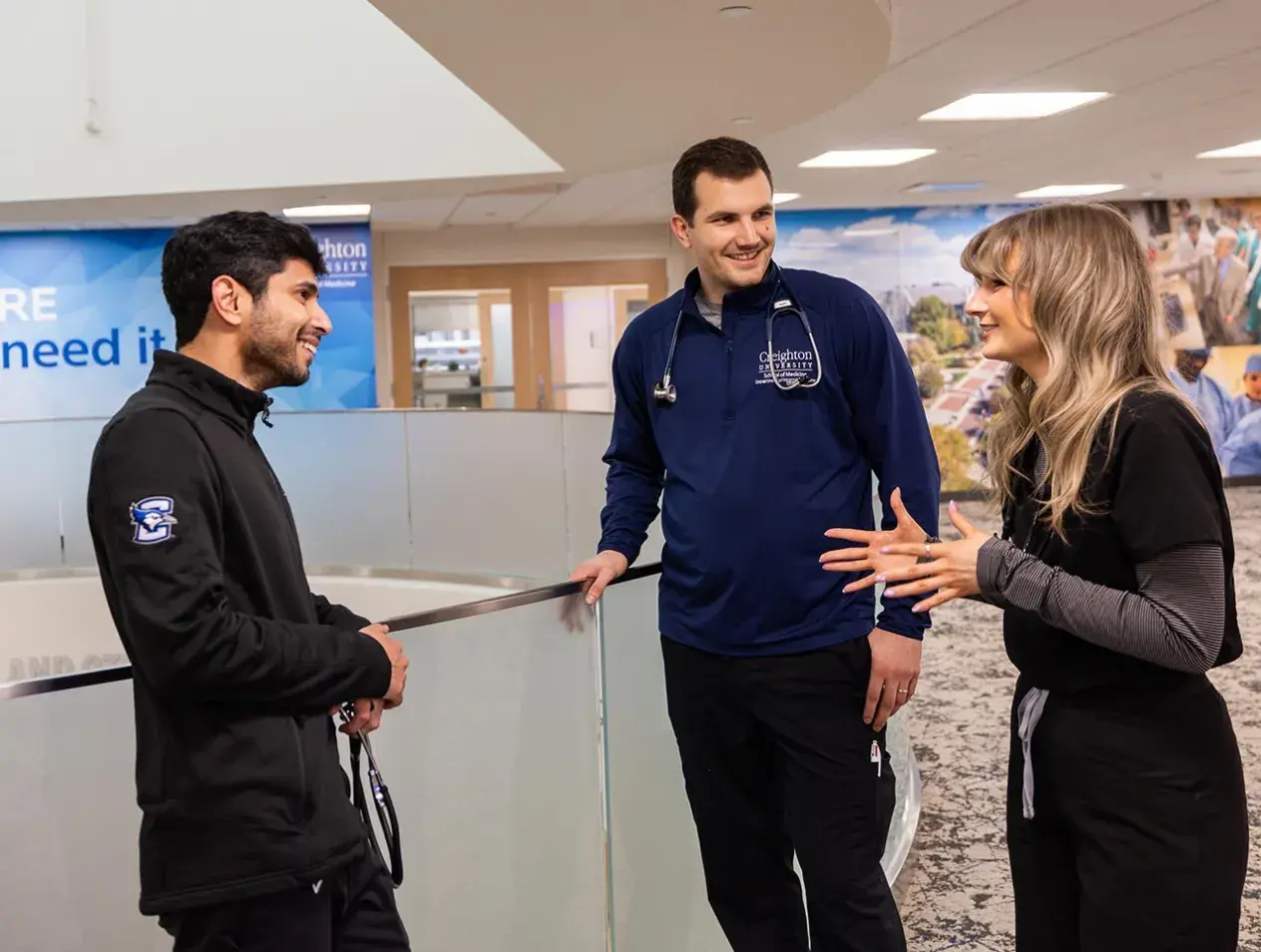 Three internal medicine residents converse in a hospital hallway in front of a 'Providing Care to Those Who Need It' banner.