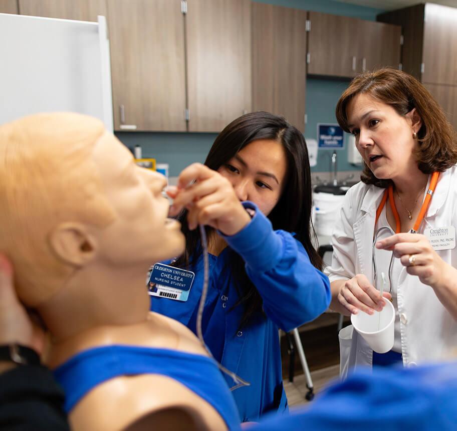 A nursing student places a tube in a practice dummy's nose while an instructor gives direction