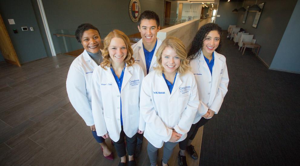 Five dentals students in white coats smiling at the camera.