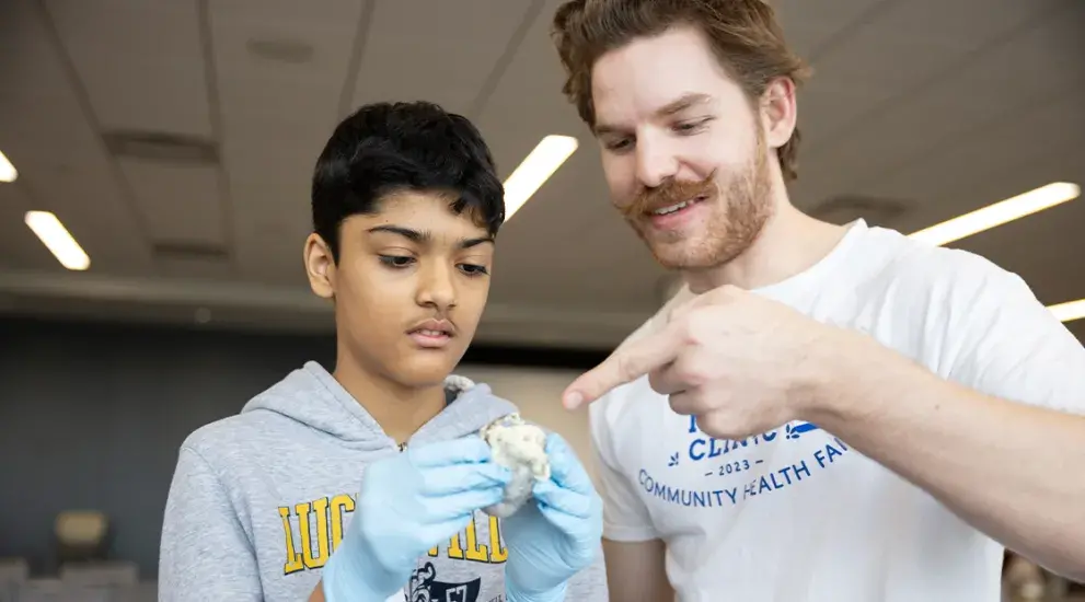 This image shows two individuals participating in a hands-on medical or science activity. The younger person on the left, wearing a gray hoodie and blue gloves, is carefully examining an object—likely a model or specimen. The adult on the right, dressed in a white t-shirt referencing a community health fair, is smiling and pointing at the object, seemingly providing guidance or instruction.