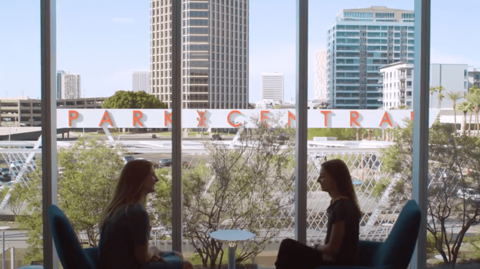 Two women facing each other in interior room with view of Phoenix downtown area seen in windows behind them.